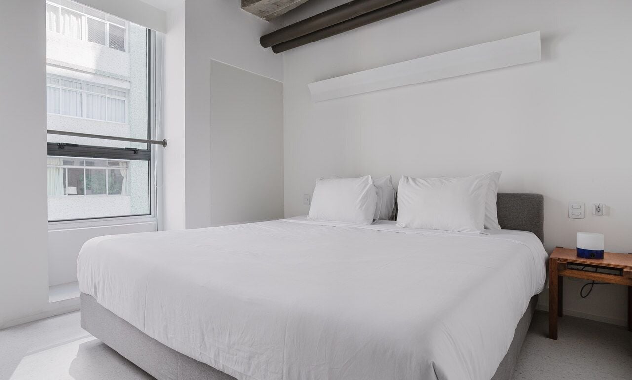 A modern bedroom with a large bed, white bedding, a wooden nightstand, and a window with blinds.
