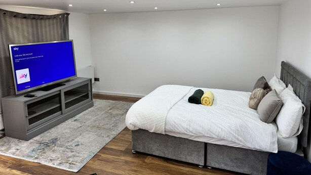 A modern bedroom with a grey sofa, a flat-screen TV displaying a Sky account login screen, a grey bed with a white duvet, and a grey headboard. - Cozy Studio - Near Stadium - Free Parking (Isleworth)
