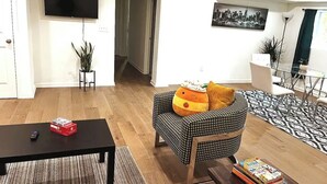 Un salón con una mesa de centro negra, un sofá a cuadros con un cojín de color calabaza, un televisor de pantalla plana y un cuadro de un paisaje urbano en la pared.