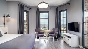 Un dormitorio moderno con una cama grande, dos sillones, una mesita redonda, un televisor y ventanas amplias con cortinas.
