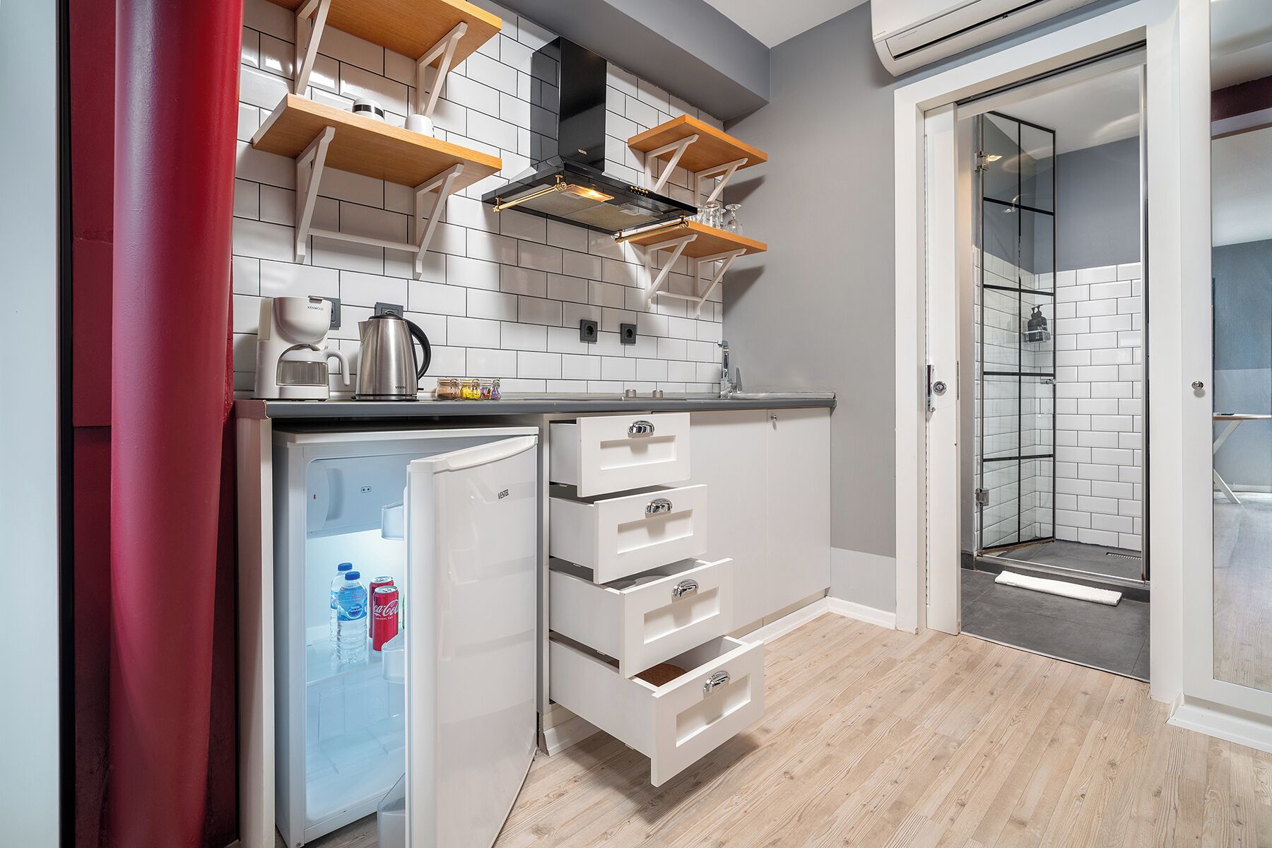 Una cucina compatta con mobili bianchi, forno integrato e frigorifero. Sopra il piano cottura c'è una mensola in legno e nel bagno adiacente si nota una cabina doccia in vetro.