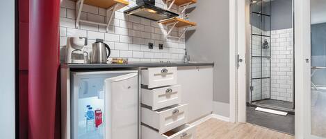 Una cucina compatta con mobili bianchi, forno integrato e frigorifero. Sopra il piano cottura c'è una mensola in legno e nel bagno adiacente si nota una cabina doccia in vetro.