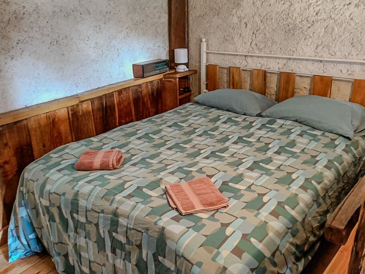 A bedroom with a wooden headboard, a patterned bedspread, two pillows, and a bedside table with a lamp and radio.