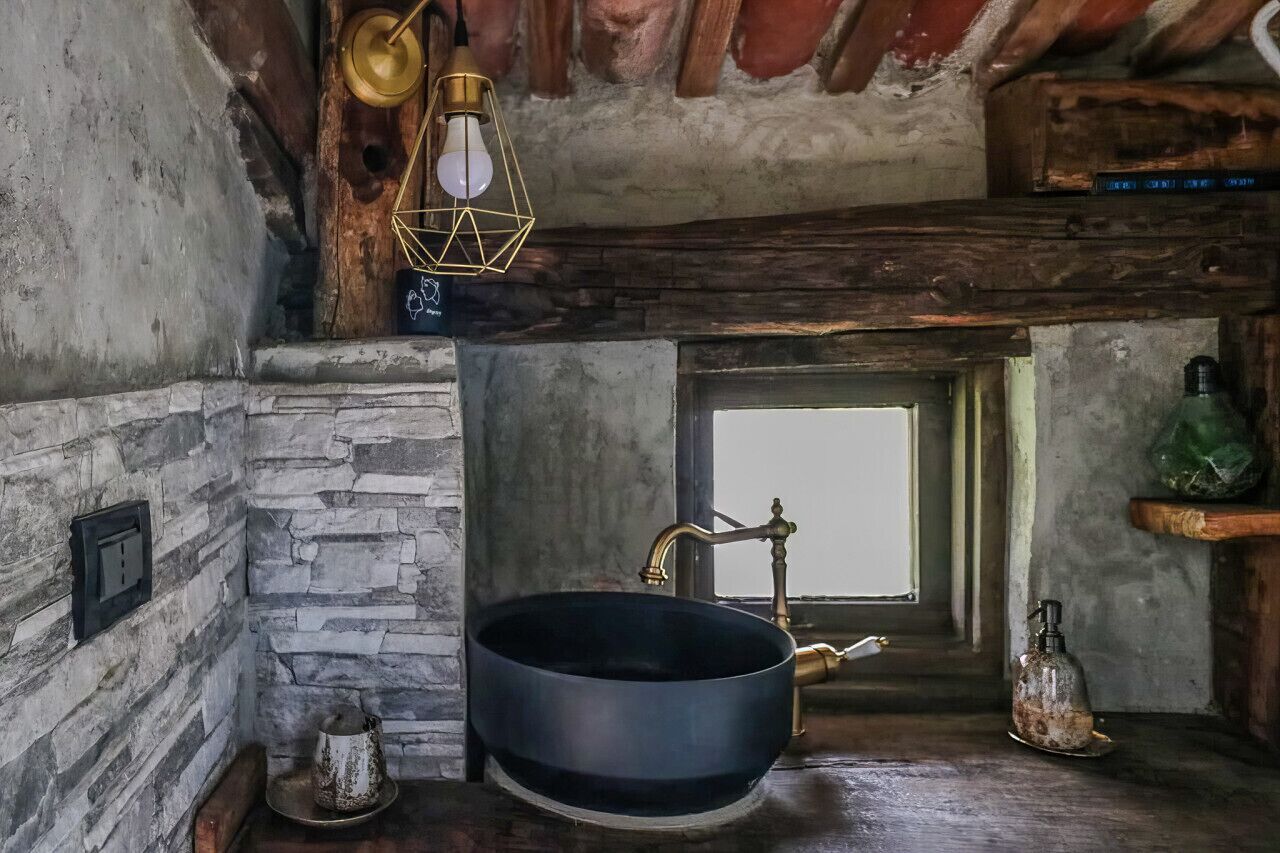 Una cocina rústica con pared de piedra, una olla negra grande sobre la encimera, un grifo antiguo, una lámpara colgante y una ventana pequeña.