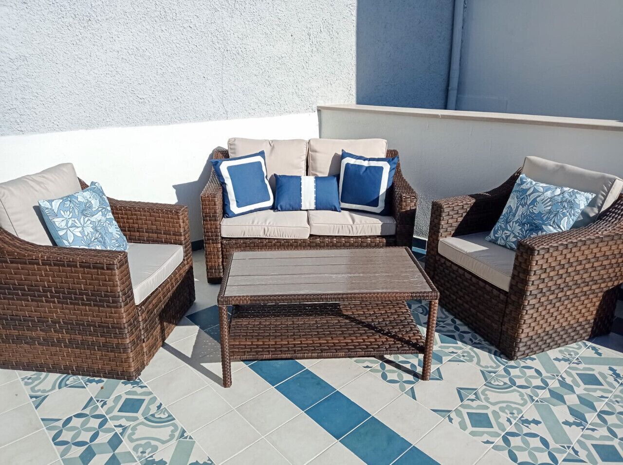 Un espacio de estar al aire libre con muebles de mimbre, una mesa, sillas acolchadas y baldosas de suelo con diseño.