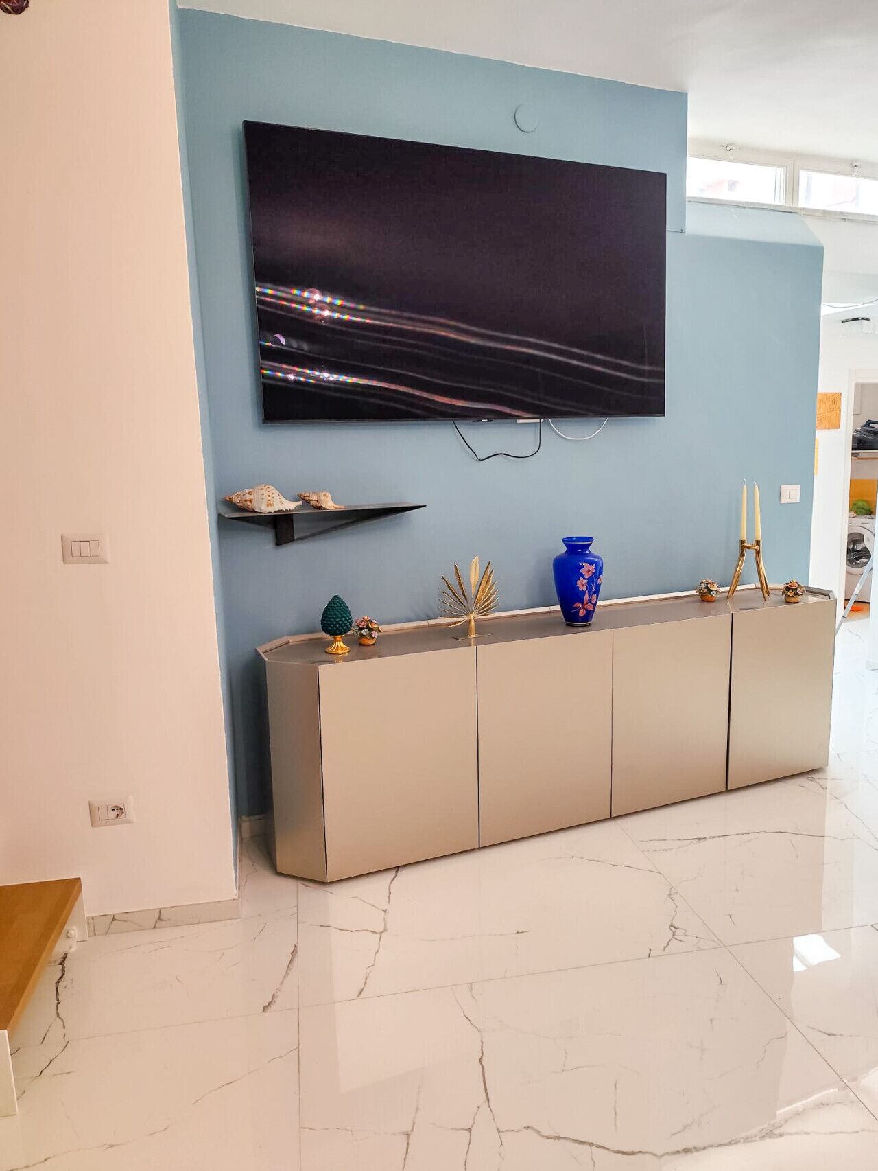A modern living room with a large flat-screen TV mounted on a light blue wall, a beige console table with decorative items, and a marble floor.