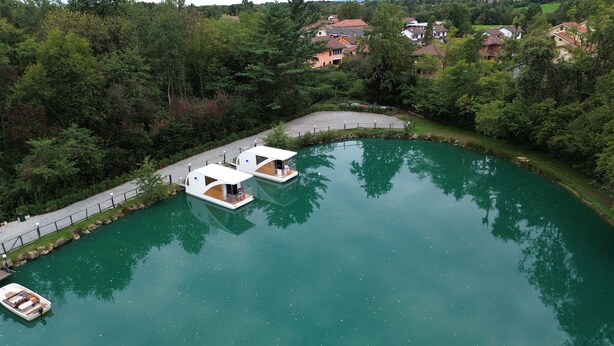 A tranquil lake with two houseboats docked, surrounded by lush greenery and residential buildings in the background. - Floating Dream, Houseboat (Fiano)