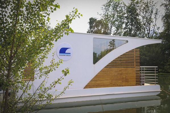 A floating house with a modern design, featuring a large window and a logo on the side. - Floating Dream, Houseboat (Fiano)