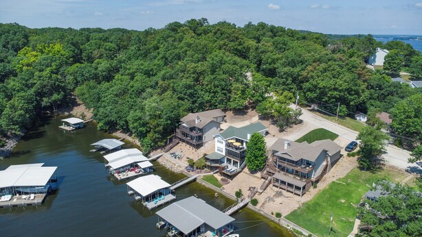 A lakeside property with multiple docked boats, a large house with a deck, and a well-maintained lawn. - Captain's Quarters, Stylish Lakefront Home, private dock with two slips, 3 bedrooms and 3 bathrooms (Lake Ozark)