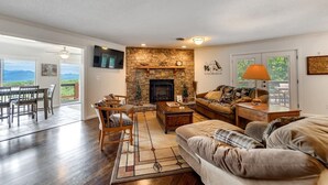 A spacious living room with a stone fireplace, wooden furniture, and a view of the outdoors. - Mountain View 4BR Home with Fireplace & Ping Pong (Franklin)