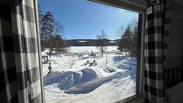 A view through a window with a checkered curtain, featuring a snowy landscape with a clear sky, trees, and a distant building.
