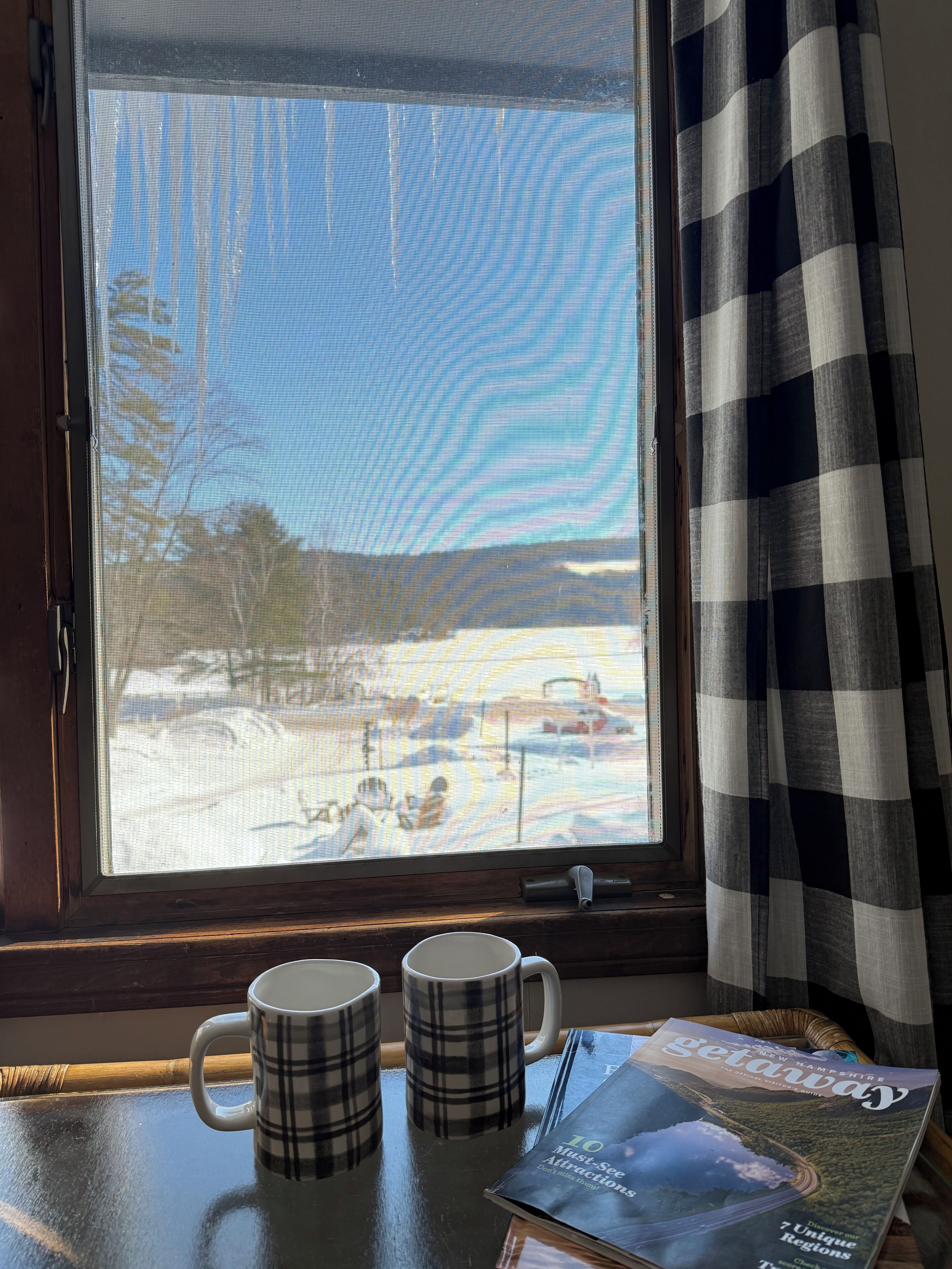 Une fenêtre donnant sur un paysage enneigé, deux tasses à motifs écossais posées sur une table et un magazine intitulé « Évasion ».