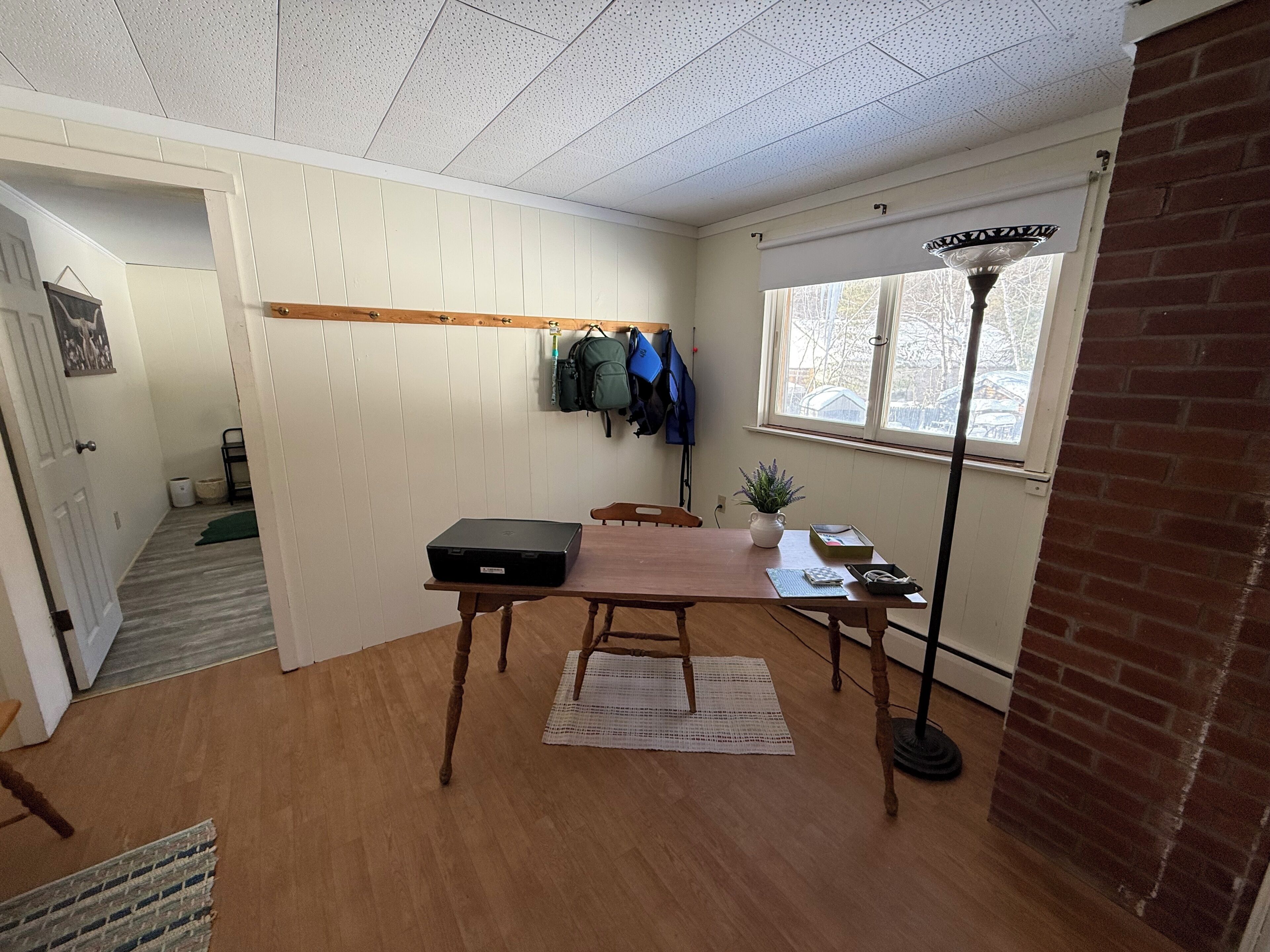 Ein Zimmer mit einem Holztisch, einem Laptop, einer Topfpflanze und einem Fenster mit Blick auf Bäume.