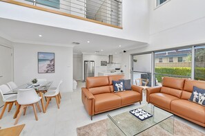 A modern apartment with a dining area, a living room with leather sofas, a kitchen, and a large window offering a view of the outdoors.