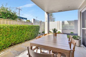 Pátio externo com mesa e cadeiras de madeira, cercado por um baixo sebedo e um muro branco.
