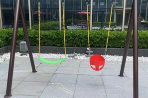 Een schommel met twee schommels, één groen en één rood, in een geplaveid buitengebied met een haag en een gebouw op de achtergrond.