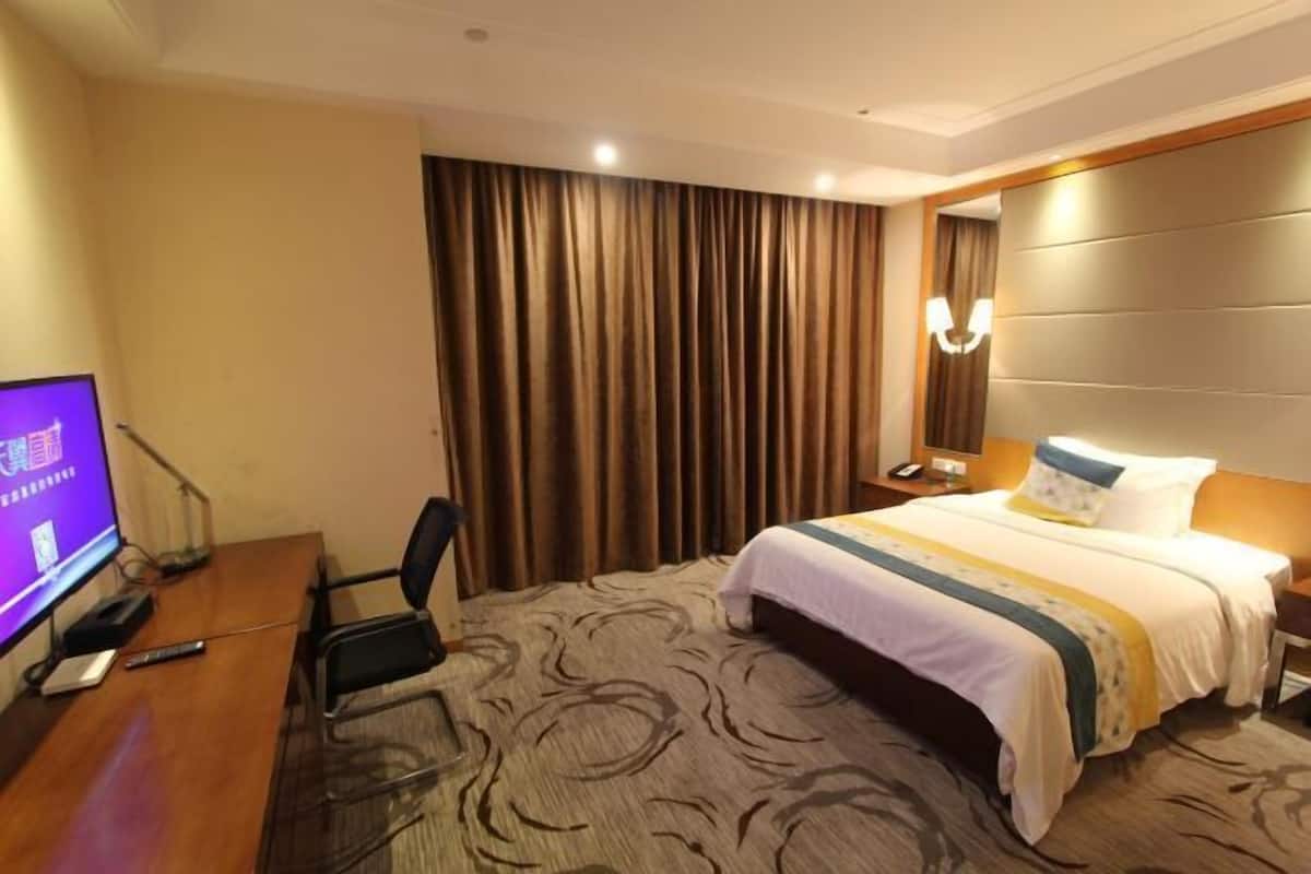 Una camera d'albergo con un letto, una scrivania con un computer, una televisione e un'ampia finestra con tende.