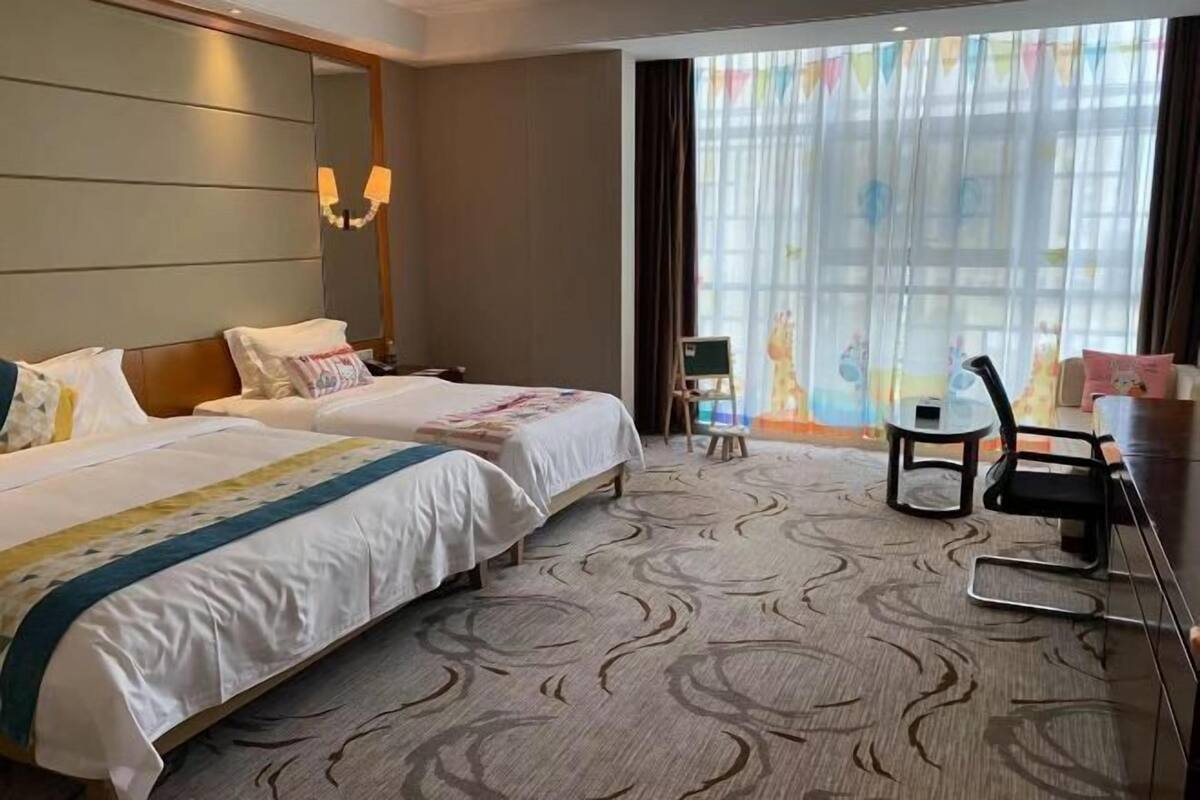 Camera d'albergo con due letti, una scrivania, una sedia, un tavolino e un'ampia finestra con tende.