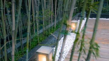 A pathway with wooden decking and stone edging, flanked by bamboo plants and illuminated lanterns.