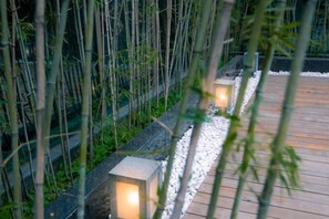Un sentier agrémenté de planches de bois et bordé de pierres, de part et d’autre duquel se dressent des plants de bambou et des lanternes illuminées.