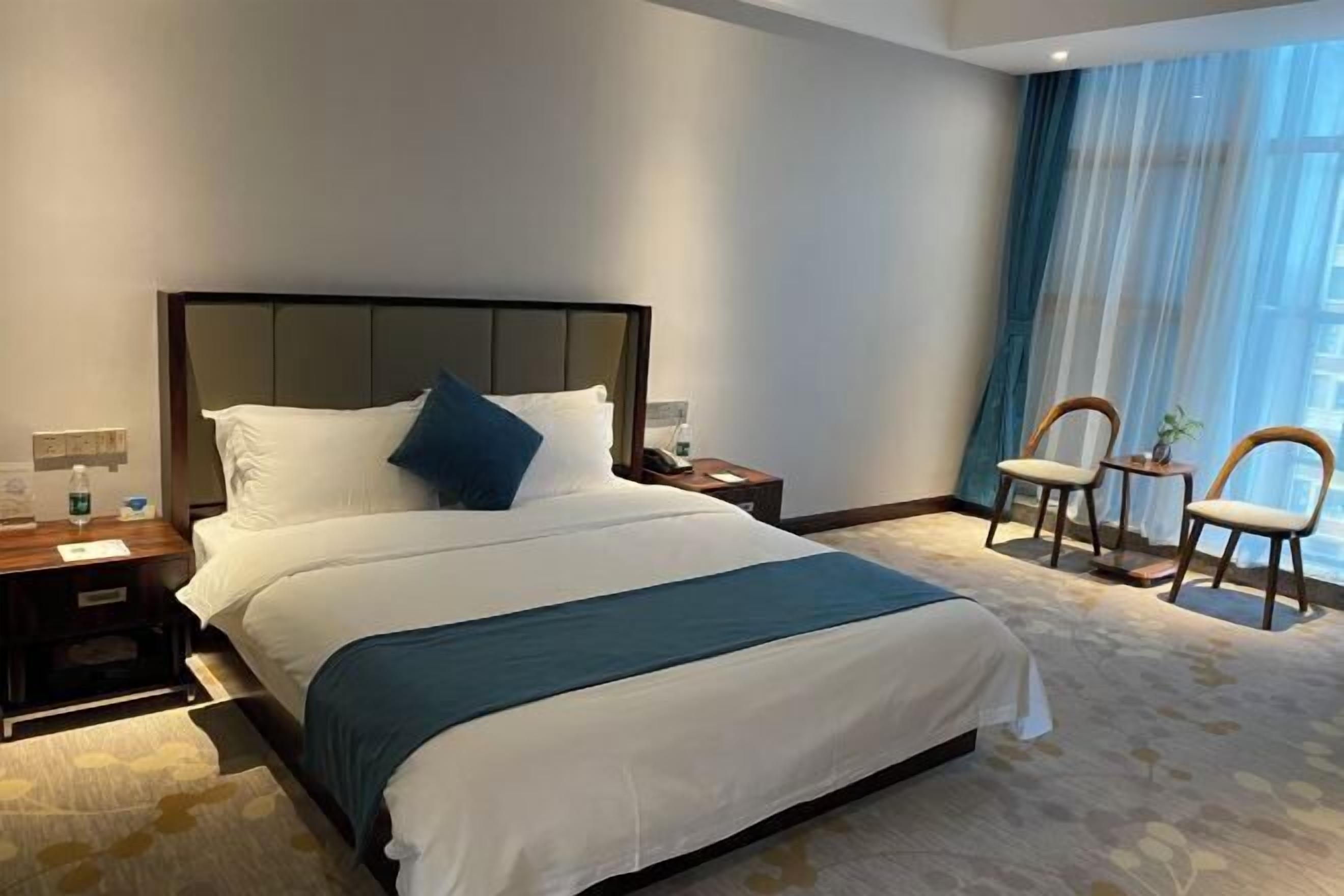 Una habitación de hotel con una cama grande, una mesita de noche, una silla y una ventana con cortinas translúcidas.