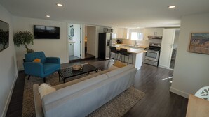 A modern living room with a sofa, armchair, and coffee table. A kitchen with white cabinets and appliances is visible in the background. - Serenity: Rockcliffe Getaway (Oliver)