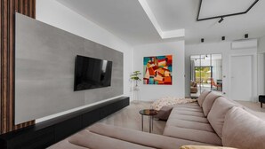 Ein modernes Wohnzimmer mit einem großen Flachbildfernseher an der Wand, einem farbenfrohen abstrakten Gemälde, einem beigefarbenen Sofa und einem kleinen Beistelltisch.