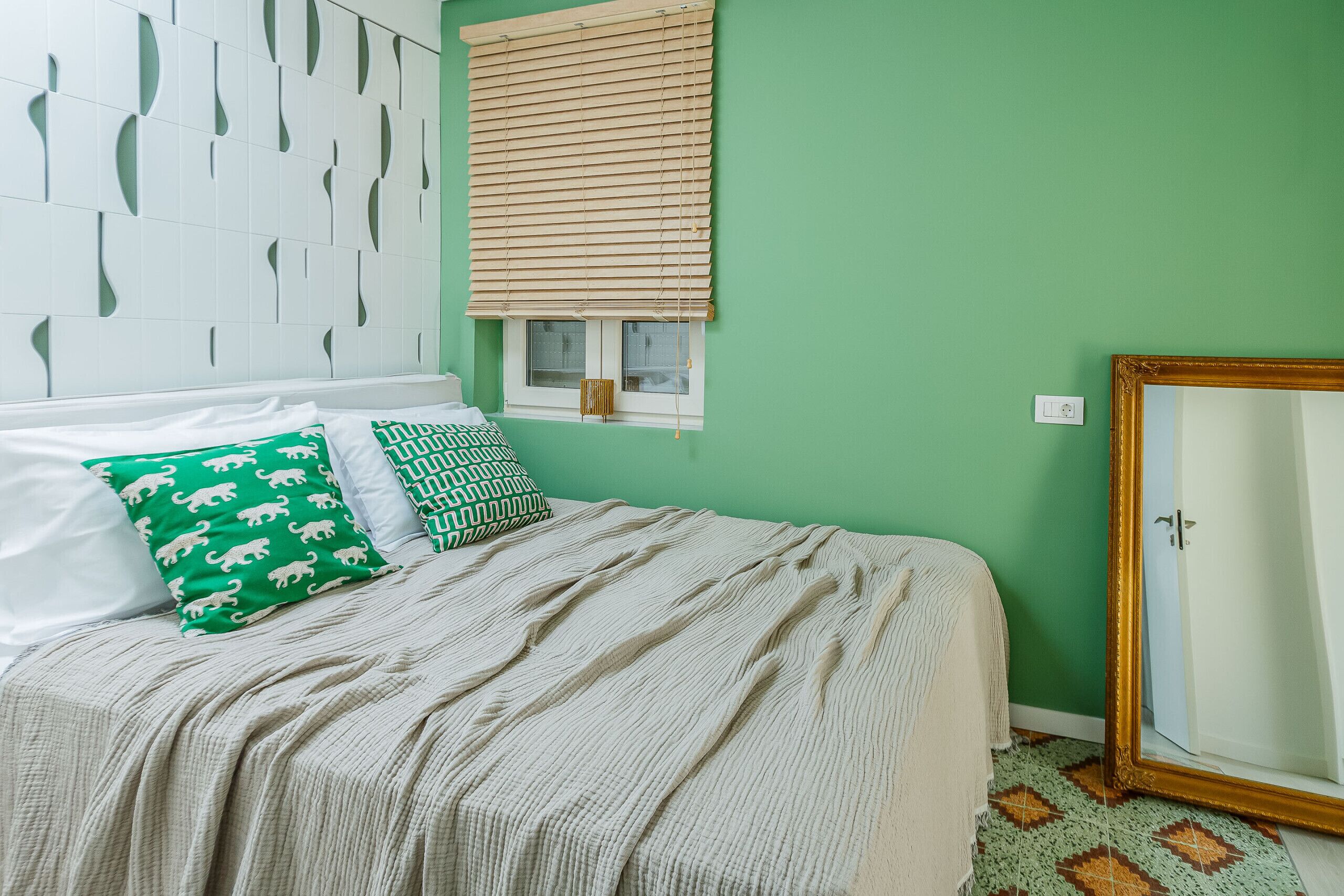 A bed with a beige blanket, two pillows with animal patterns, a window with blinds, and a mirror with a gold frame.
