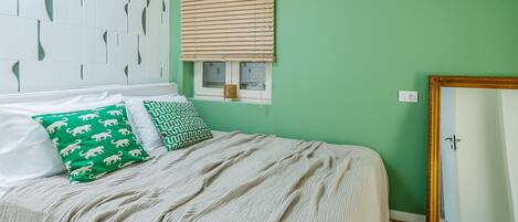A bed with a beige blanket, two pillows with animal patterns, a window with blinds, and a mirror with a gold frame.