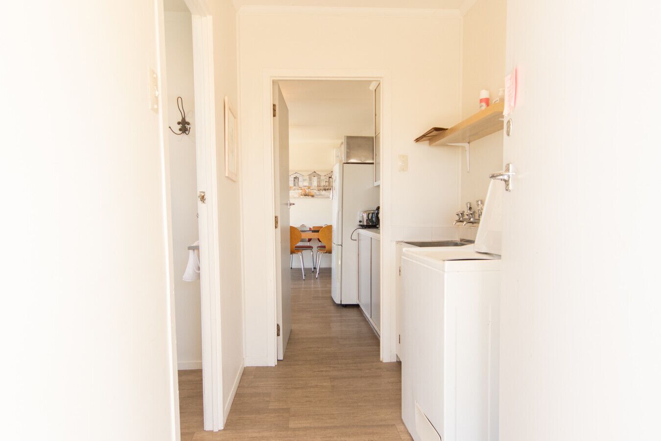 A narrow hallway leading to a kitchen with a washing machine, a refrigerator, and a dining area with chairs and a table.