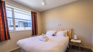 Una cama bien hecha con sábanas blancas y una toalla enrollada, una mesita de noche con lámpara, y una ventana con cortinas.