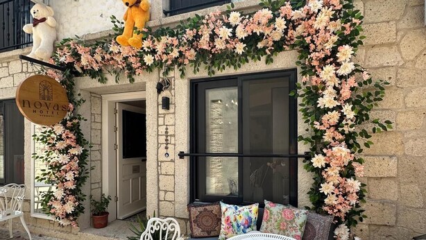 A hotel exterior with a stone facade, a balcony with two teddy bears, a floral arch, and a seating area with white chairs and a round table. - Novas Hotel Alaçatı (Cesme)