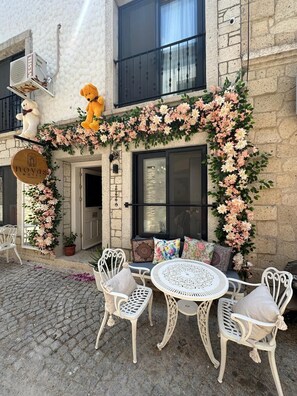 A hotel exterior with a stone facade, a balcony with two teddy bears, a floral arch, and a seating area with white chairs and a round table. - Novas Hotel Alaçatı (Cesme)