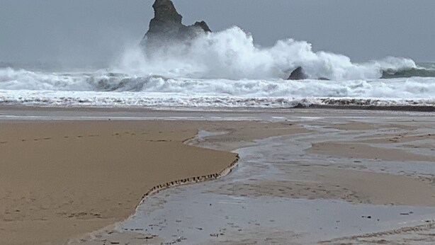 Ein Strand mit brechenden Wellen und einer markanten Felsformation im Hintergrund.