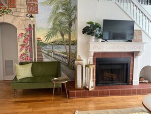 Ein Wohnzimmer mit einem grünen Sofa, einem Kamin, einem Fernseher und einem Wandbild mit einer Brücke und Palmen.