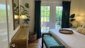 A bedroom with a bed, bedside lamps, a mirror, a wooden dresser, a bench, and a view of the outdoors through a door. - Charming 2-bedroom cottage in downtown Mount Dora with RV parking, pets welcome  (Mount Dora)
