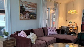 A living room with a brown sofa, a glass coffee table, a painting on the wall, and a window with curtains. - Charming 2-bedroom cottage in downtown Mount Dora with RV parking, pets welcome  (Mount Dora)