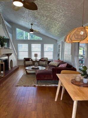 A spacious living area with a fireplace, a sofa, a coffee table, and a dining table. There are large windows allowing natural light and a view of the outdoors. - Casita Encanta-Dora, Steps to town, Pet-Friendly (Mount Dora)