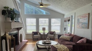 A spacious living room with a fireplace, a ceiling fan, and large windows with blinds. - Charming 2-bedroom cottage in downtown Mount Dora with RV parking, pets welcome  (Mount Dora)
