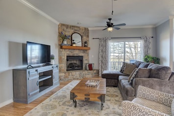 This living room combines comfort and style with its cushy couches adorned with throw pillows, a vintage center table, and a wooden cabinet housing a large TV. The fireplace with elegant accents and the large glass window with floral curtains complete the