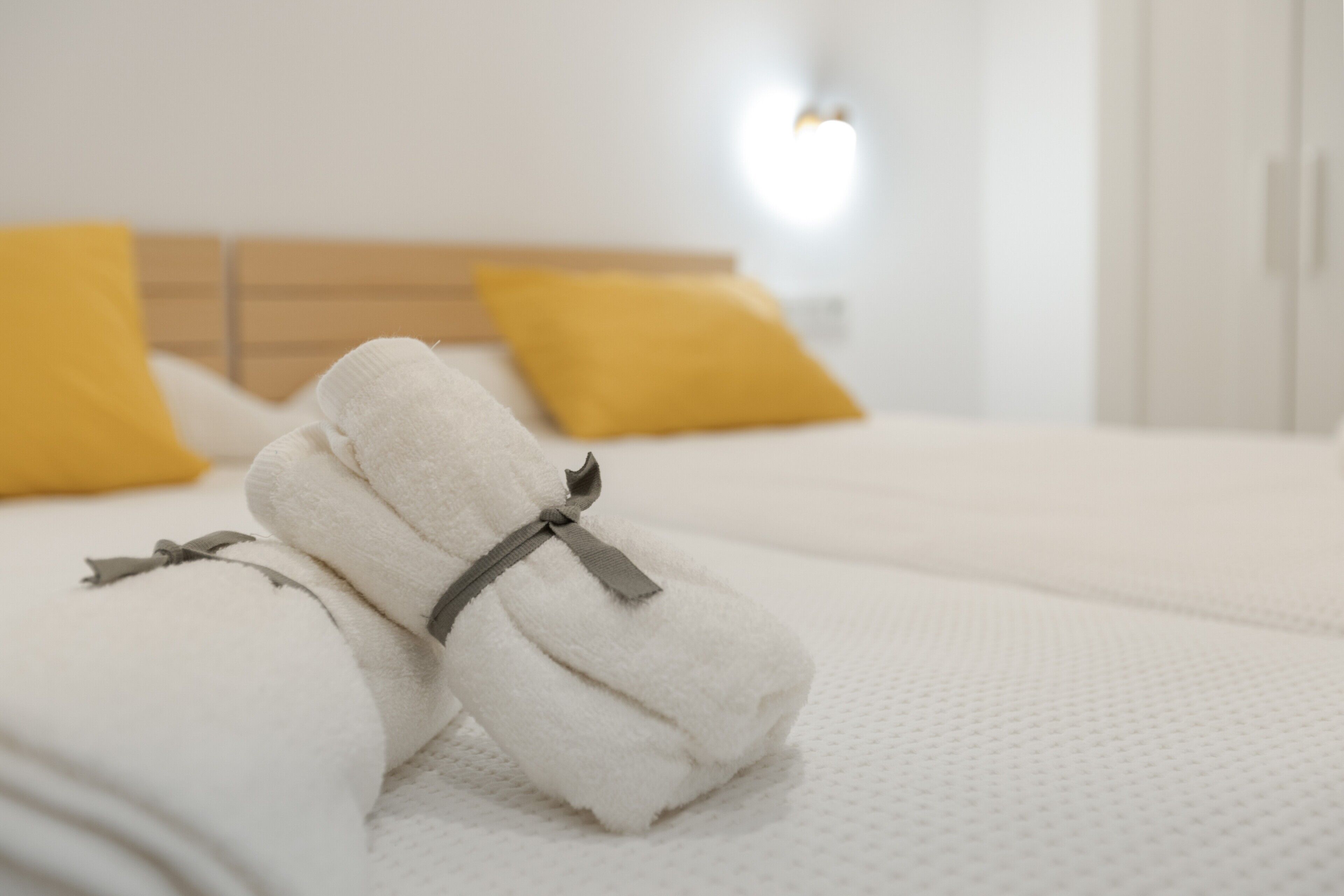 A neatly made bed with white linens and a gray ribbon, two yellow pillows, and a wooden headboard.