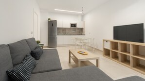 Un salon moderne avec un canapé gris, une table basse en bois, un téléviseur posé sur un meuble en bois, et une cuisine en arrière-plan.