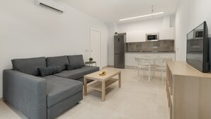Un salon moderne avec un canapé gris, une table basse en bois et un coin cuisine doté de meubles blancs et d’appareils électroménagers.
