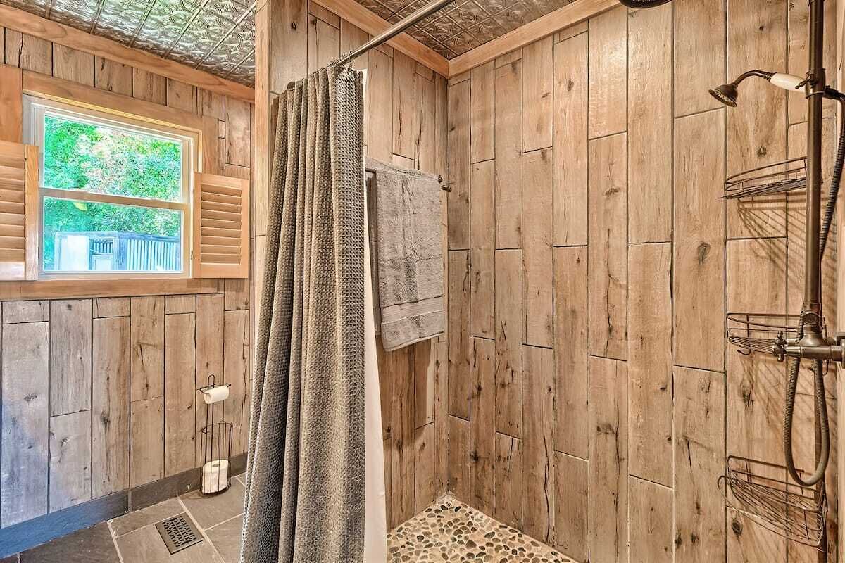 A wooden-paneled bathroom with a shower, a window with a view of greenery, and a towel rack.