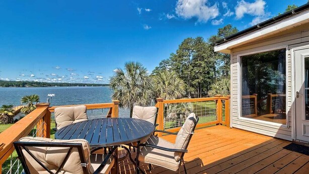 A wooden deck with a round table and chairs, overlooking a lake with trees and a clear sky. - Laid-Back River Shack (Camden)