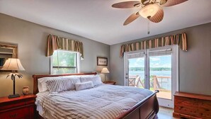 A bedroom with a large bed, two bedside lamps, a wooden nightstand, a ceiling fan, and a view of a balcony with a railing. - Laid-Back River Shack (Camden)