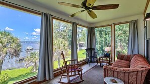 A room with a large window overlooking a body of water, a ceiling fan, a rocking chair, a sofa, and a small table. - Laid-Back River Shack (Camden)