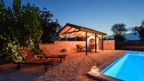 Un área junto a la piscina con un patio de piedra, sillas de descanso, una zona de estar cubierta y una piscina.