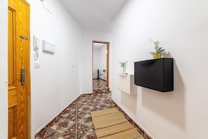 Ein schmaler Flur mit gemusterten Bodenfliesen, einer Holztür, einem an der Wand befestigten Telefon und einem schwarzen Regal mit einer Pflanze.