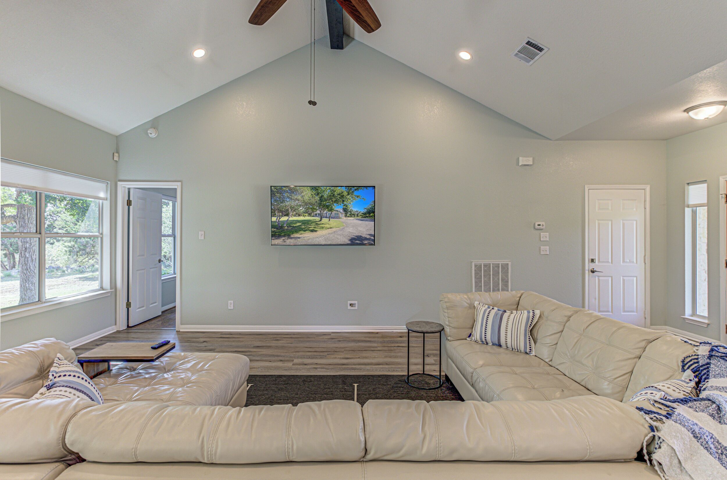 A spacious living room with a large sectional sofa, a flat-screen TV mounted on the wall, and a ceiling fan.